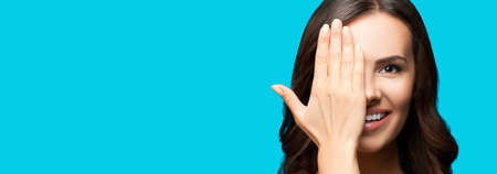Portrait studio photo of happy smiling brunette woman with one eye, closed by hand, covering part of her face, isolated over aqua blue green color background. Copy space area for text. Wide image.の写真素材