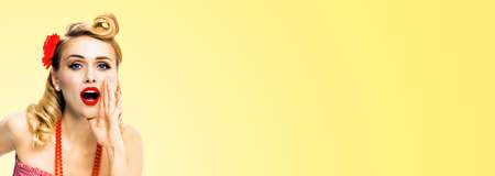 Portrait of woman holding hand near open mouth and saying something. Girl dressed in pin up style. Blond haired model at retro fashion vintage studio concept, isolated over light yellow backgroundの写真素材