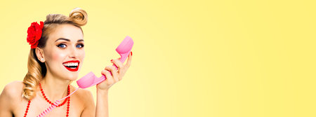 Portrait photo of happy smiling woman holding phone tube. Excited pin up girl. Beautiful model at retro fashion and vintage studio concept. Light yellow background. Copy space for text.の写真素材