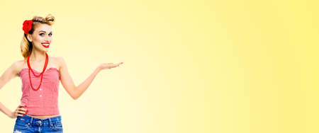 Happy excited smiling woman holding, giving, advertising something. Blond girl in pinup style, showing copyspace for text. Retro fashion and vintage. Light yellow background. Wide image.の写真素材