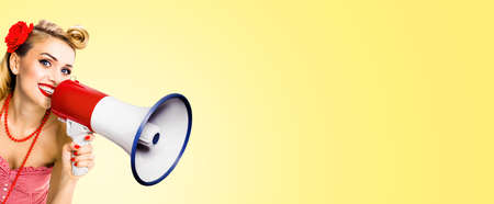 Photo of happy excited woman holding mega phone and saying something. Blond girl in pin up style with megaphone, on light yellow background with copy space area. Female model in retro vintage concept.の写真素材