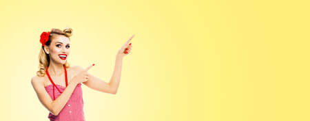 Portrait photo - happy excited smiling woman pointing something. Blond girl in pin up style, showing copy space for text or imaginary. Retro fashion and vintage. Light yellow background. Wide image.の写真素材