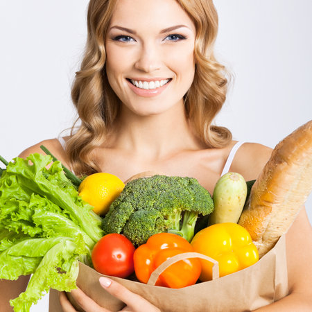 Portrait photo of happy smiling young beautiful woman holding grocery shopping bag with healthy vegetarian raw food, isolated over gray background. Attractive blond girl at studio.の写真素材