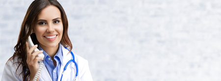 Portrait image of happy smiling female doctor on phone, over white brick loft wall background. Medical call center concept picture.の写真素材