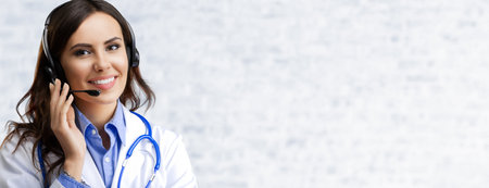 Portrait image of happy smiling female doctor in headset, over white brick loft wall background, with empty copy space area. Medical call center concept picture.の写真素材