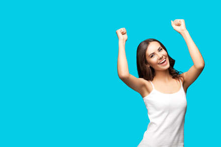 Photo of happy gesturing beautiful woman rising hands up with clenching fists, in white smart casual cloth, isolated on aqua blue green color background. Excited brunette girl at studio.の写真素材