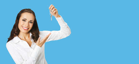 Happy smiling young businesswoman or real estate agent showing keys from new house. Portrait of brunette woman at studio, bright blue color background with copy space area. Wide composition.の写真素材