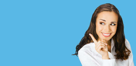 Young happy smiling woman in white confident blouse, showing pointing at copy space for text or imaginary. Business ad concept. Bright blue color background. Brunette businesswoman. Wide.の写真素材