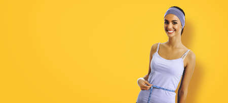 Woman in sportswear measuring waist with a tape measure, over yellow background. Sporty african american girl at studio. Health, beauty and fitness gym center concept.の写真素材