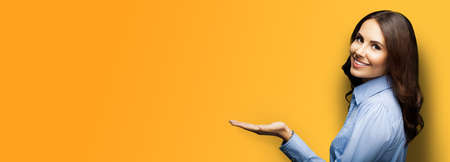 Happy smiling young businesswoman holding or giving something. Brunette girl showing copy space for some text, isolated over orange yellow color background. Wide banner composition image.の写真素材