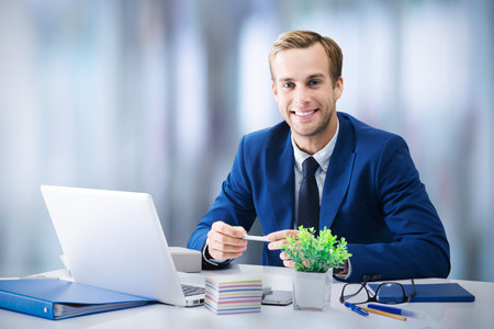 Young happy smiling businessman in blue suit working with laptop computer at modern office. Success...