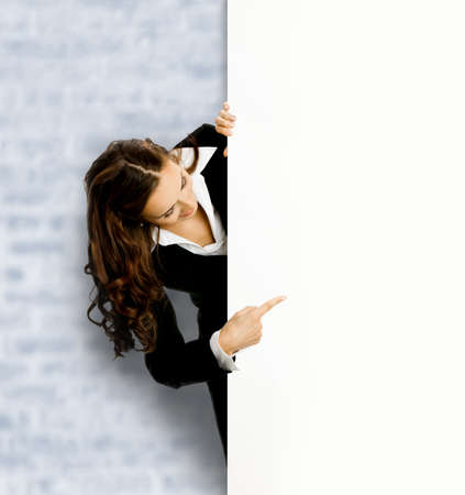 Happy smiling business woman, standing behind, peeping from blank banner or mock up signboard, showing pointing copy space, on white bricks loft wall backgroundの写真素材