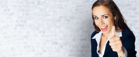 Businesswoman in confident black suit, showing thumbs up gesture, copy space area. Business concept. White bricks loft wall background. Brunette businesswoman, indoors.の写真素材