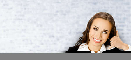 Businesswoman in black suit showing call me hand sign gesture, on white bricks loft wall background. Portrait of happy smiling gesturing brunette woman. Business concept.の写真素材