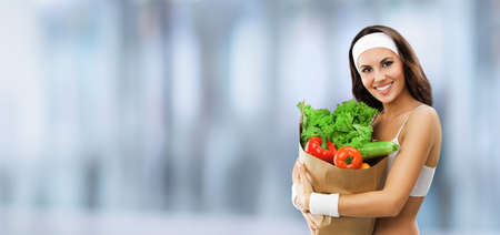 Portrait image of happy smiling young beautiful brunette woman in fitness wear holding grocery shopping bag with vegetarian raw food, vegetables, inside. Healthy eating, dieting concept.の写真素材