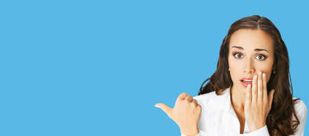 Portrait image of excited surprised, shocked astonished businesswoman pointing away side with finger. Business woman covering open mouth by her hand. Blue color background. Wide banner composition.の写真素材