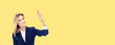 Smiling young businesswoman in confident suit, writing or drawing something on screen or transparent glass, by marker, isolated over yellow background.の写真素材