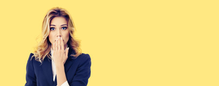 Shocked businesswoman in dark blue confident suit, with wide open eyes, covering mouth by her hand, on yellow color background. Blond woman in business studio concept.の写真素材