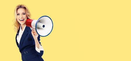 Happy excited young business woman in suit, holding megaphone and shout something. Confident businesswoman in advertising studio concept, isolated over yellow color background. Copy space.の写真素材