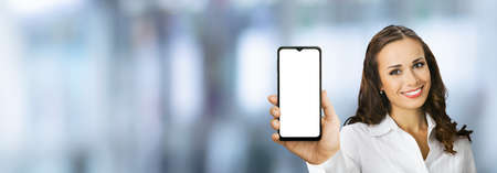 Portrait image of happy smiling young woman in confident white clothing holding showing smartphone cell phone mobile with empty mock up screen, over blurred office background. Consulting assistance.の写真素材