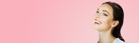 Portrait of happy excited smiling beautiful young woman with open mouth, looking side away up, isolated over rose pink background. Brunette laughing girl at studio image. Wide banner composition.の写真素材