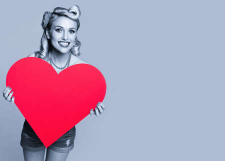 Portrait of beautiful happy smiling woman holding heart symbol, dressed in pin up style. Blond girl posing in retro fashion and vintage concept. Black and white. Copy space.の写真素材
