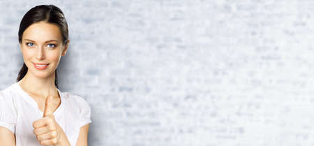 Portrait image of brunette businesswoman showing thumb up gesture, copy space place for ad text, advertising or slogan. Young business woman, over white bricks wall.の写真素材