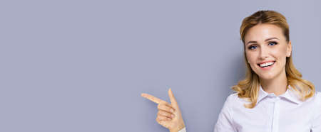 Young confident woman, showing pointing finger at something or copy space area for text. Success in business ad concept. Grey color background. Wide horizontal banner composition.の写真素材