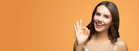 Photo of woman making ok okay hand sign or zero gesture, isolated over vivid brown color background. Happy smiling gesturing brunette girl at studio. Wide horizontal banner composition.の写真素材