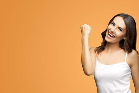 Photo of happy gesturing beautiful woman with open mouth raising hand up with clenching fist, in white smart casual clothing, isolated over vivid brown background. Excited brunette girl at studio.の写真素材
