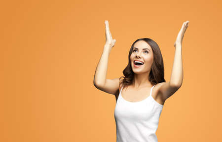 Photo of woman in white smart casual clothing raising hands up with flat palms, happy gesturing or praying, isolated on vivid brown background. Excited brunette girl looking up with smile, at studio.の写真素材