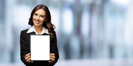 Portrait of smiling brunette businesswoman showing tablet pc, touchpad, with copy space, over blurred office interior background. Confident beautiful young business woman indoors.の写真素材