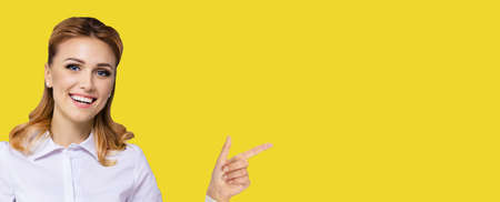 Young attractive confident woman, showing pointing finger at something or copy space empty text area. Success in business ad concept. Yellow color background. Wide horizontal banner composition.の写真素材