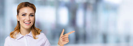 Young attractive confident woman, showing pointing finger at something or copy space empty text area. Success in business ad concept. Blurred office background. Wide horizontal banner composition.の写真素材