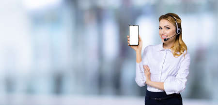 Customer support phone operator in headset holding showing smartphone cell phone mobile with white mock up screen, over blurred office background. Consulting and assistance service call center.の写真素材