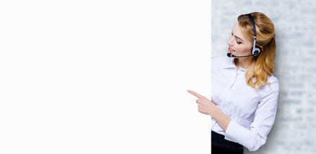 Call Center Service concept - customer support or sales agent. Businesswoman, caller, phone operator pointing finger at empty white signboard. Helping, answering, consulting. White bricks wall.の写真素材