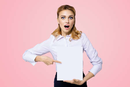 Very happy, excited surprised, astonished businesswoman with wide opened eyes, mouth, showing blank paper signboard. Success in business concept. Copy space place for text. Rose pink color background.の写真素材