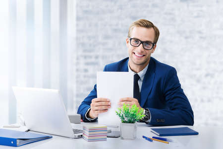 Young confident businessman in glasses and blue suit working with documents and laptop computer at office workplace. Portrait of happy man - business, job and education concept image.の写真素材