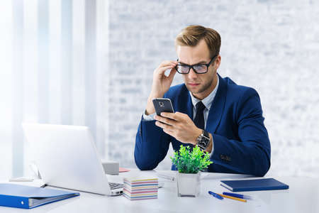 Serious businessman in glasses and blue suit working with cellphone and laptop computer at office. Success in business, job and education concept.の写真素材