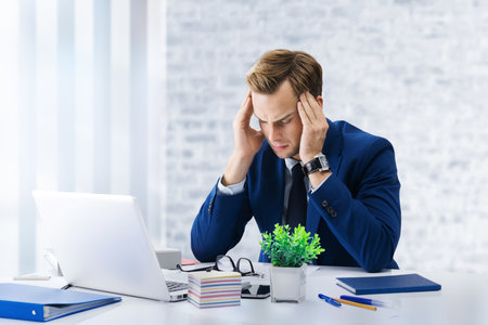 Stressed, tired businessman or heaving headache, holding his head, with laptop computer at office workplace. Portrait of confident man - business, job and education concept image.の写真素材