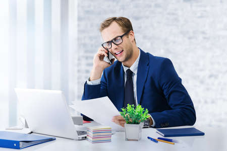 Young happy smiling businessman in glasses and blue suit working with cellphone and laptop computer at office  workplace. Portrait of happy man - business, job and education concept image.の写真素材