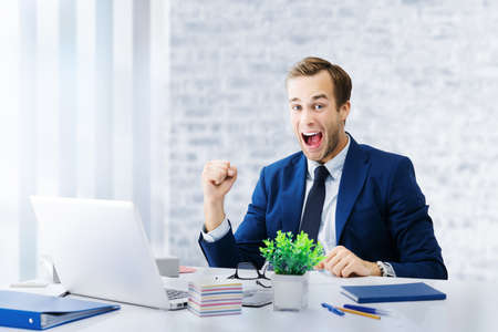 Great deal! Very happy businessman in blue suit, working with laptop computer at office. Excited business man with rised hand up with clenching fist - success in job and education concept.の写真素材