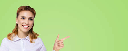 Young attractive confident woman, showing pointing finger at something or copy space empty text area. Success in business ad concept. Green colour background. Wide horizontal banner compositionの写真素材