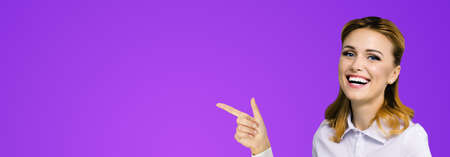 Young attractive confident woman, showing pointing finger at something or copy space empty text area. Success in business ad concept. Businesswoman on violet purple. Wide horizontal banner compositionの写真素材