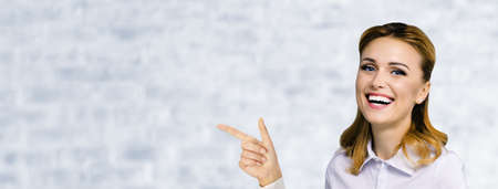 Young attractive confident woman, showing pointing finger at something or copy space empty text area. Success in business ad concept. White bricks wall. Wide horizontal banner compositionの写真素材