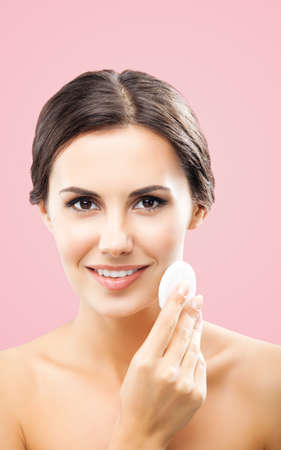 Portrait of beautiful dark haired woman cleaning face skin by cotton pad, isolated over rose pink background. Happy smiling brunette girl at studio. Beauty model picture.の写真素材