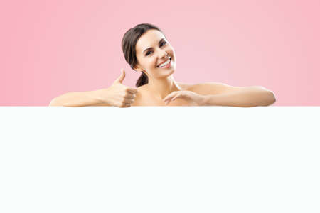 Excited smiling brunette woman with naked shoulders, undress, showing thump up hand sign gesture, standing behind white banner mock up signboard with copy space area, over rose pink color background.の写真素材