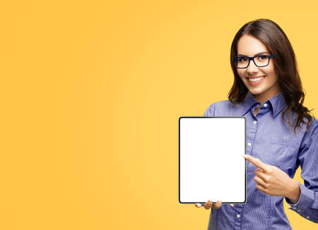 Happy smiling businesswoman in eye glasses spectacles showing touchpad tablet pc, with white screen copy space text area for advertising or slogan sign, isolated over yellow background. Business womanの写真素材