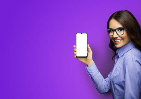 Portrait image of happy smiling young woman in eye glasses holding showing smartphone cell phone mobile with white mock up screen, isolated on purple color background. Consulting assistance.の写真素材