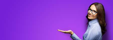 Happy smiling businesswoman holding or giving something. Brunette girl in glasses showing copy space for text or product, isolated on purple background. Wide banner composition image.の写真素材